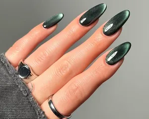 Hand displaying long almond-shaped green nails with a shiny velvet finish, silver rings on the fingers and dark clothing visible