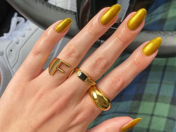 A hand with long manicured nails wearing gold rings nails adorned with shiny golden polish